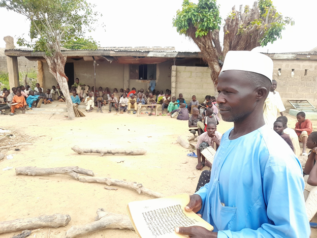Right; Mallam Ibrahim Hassan standing, while the Almajirai studying under scorching sun. PC: Lukman Abdulmalik.